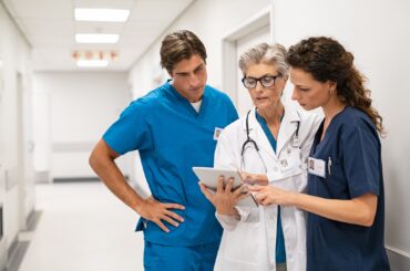 Mature female doctor discussing medical report with nurses in hospital hallway. Senior general practitioner discussing patient case status with group of medical staff after surgery. Doctor working on digital tablet while in conversation with healthcare workers, copy space.