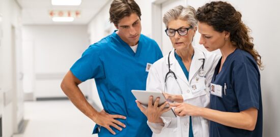 Mature female doctor discussing medical report with nurses in hospital hallway. Senior general practitioner discussing patient case status with group of medical staff after surgery. Doctor working on digital tablet while in conversation with healthcare workers, copy space.